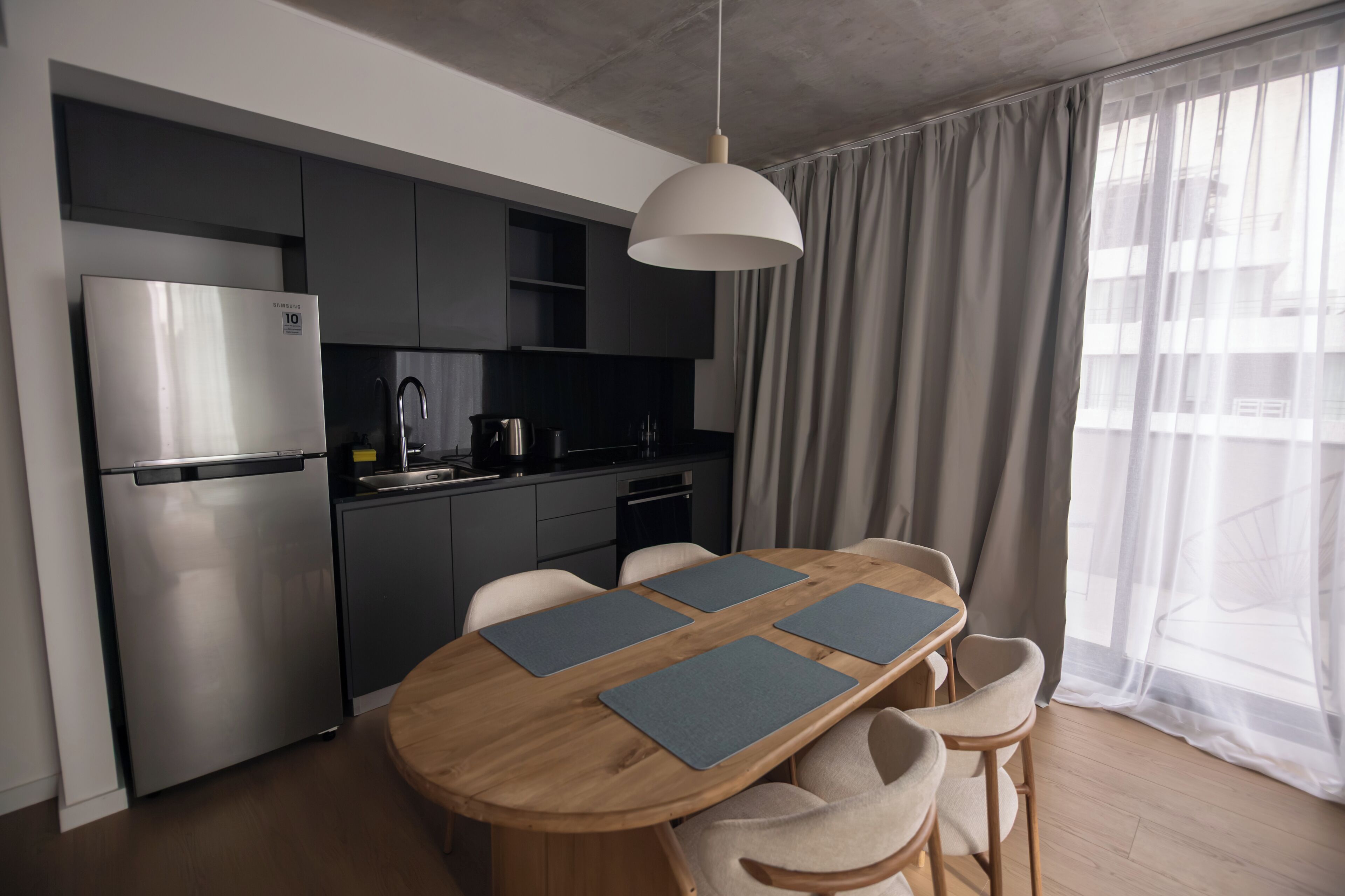 Une cuisine moderne équipée d’un réfrigérateur en acier inoxydable, d’un four encastré et d’une machine à café. On y trouve une table à manger en bois avec quatre chaises, et un luminaire suspendu au-dessus.