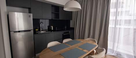 A modern kitchen with a stainless steel refrigerator, a built-in oven, and a coffee maker. There is a wooden dining table with four chairs and a hanging light fixture above it.