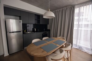 Una cocina moderna con un refrigerador de acero inoxidable, un horno empotrado y una cafetera. Hay un comedor de madera con cuatro sillas y una lámpara colgante arriba.
