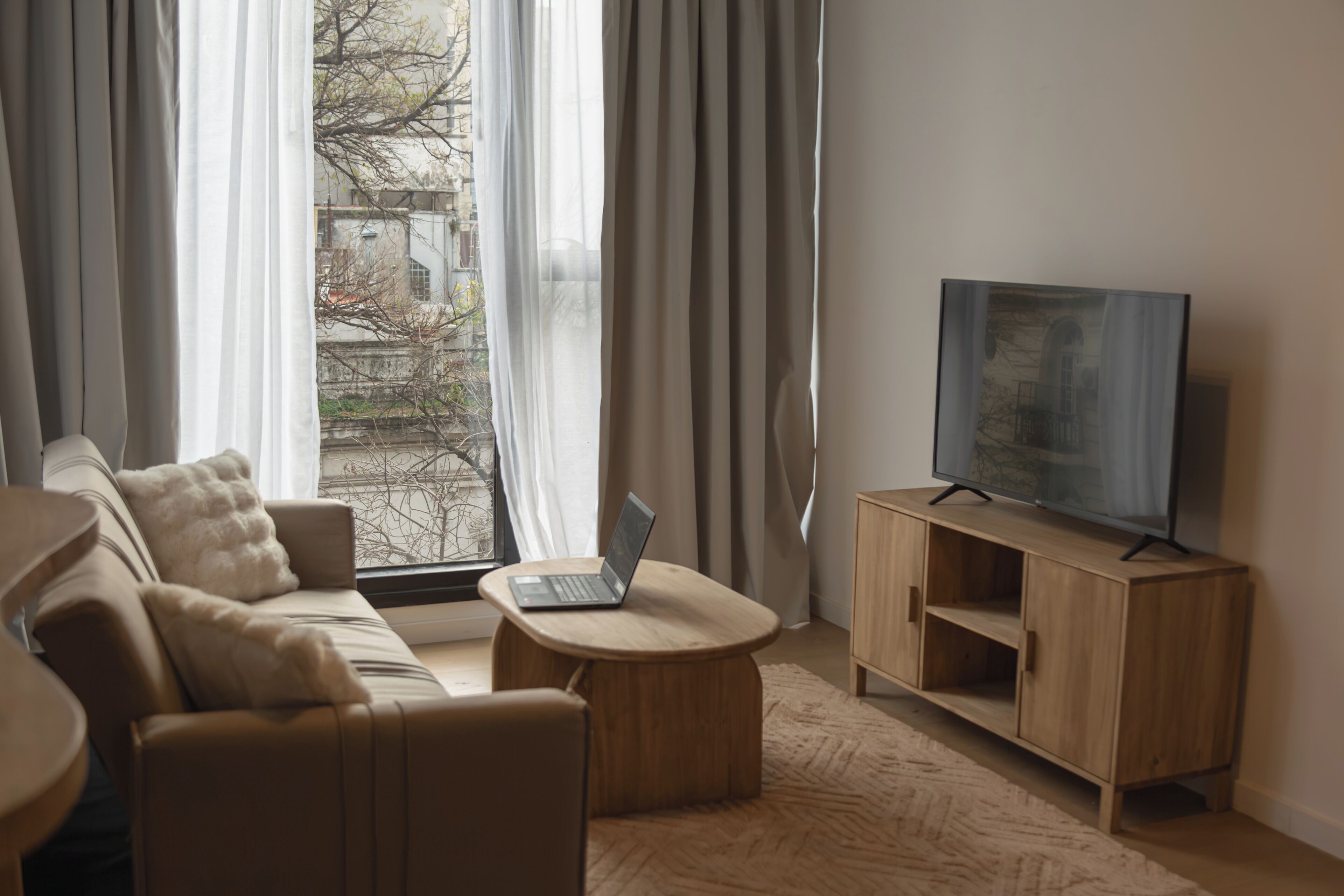 Una sala de estar moderna con sofá, una mesa de centro, un televisor sobre un mueble de madera y una ventana con cortinas.
