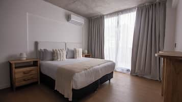 Un dormitorio con cama, mesitas de noche de madera, una mesilla auxiliar, una ventana con cortinas y un equipo de aire acondicionado en la pared.