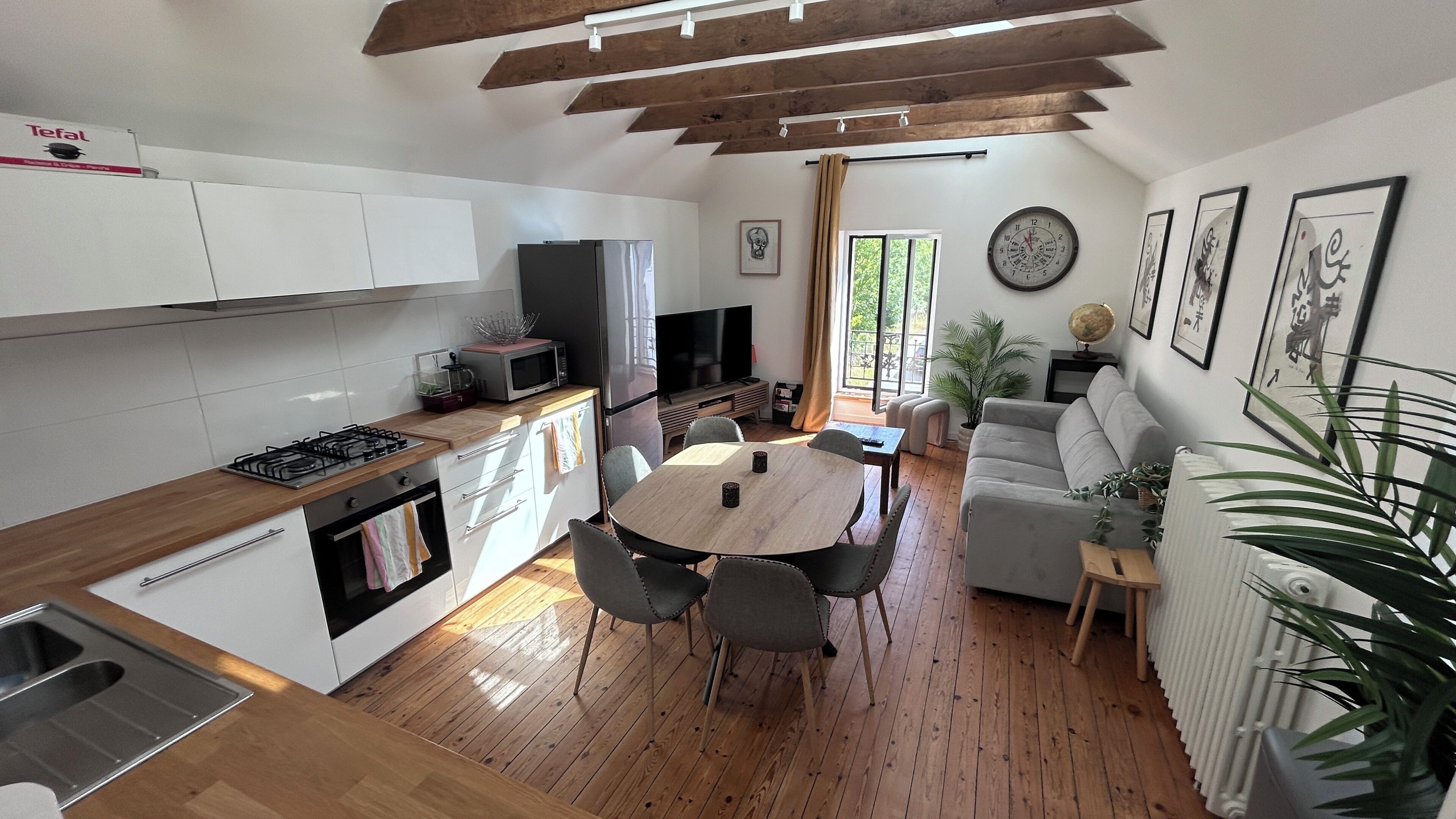 Une cuisine moderne avec des meubles blancs, une table à manger avec des chaises, un canapé, un téléviseur et une horloge au mur.