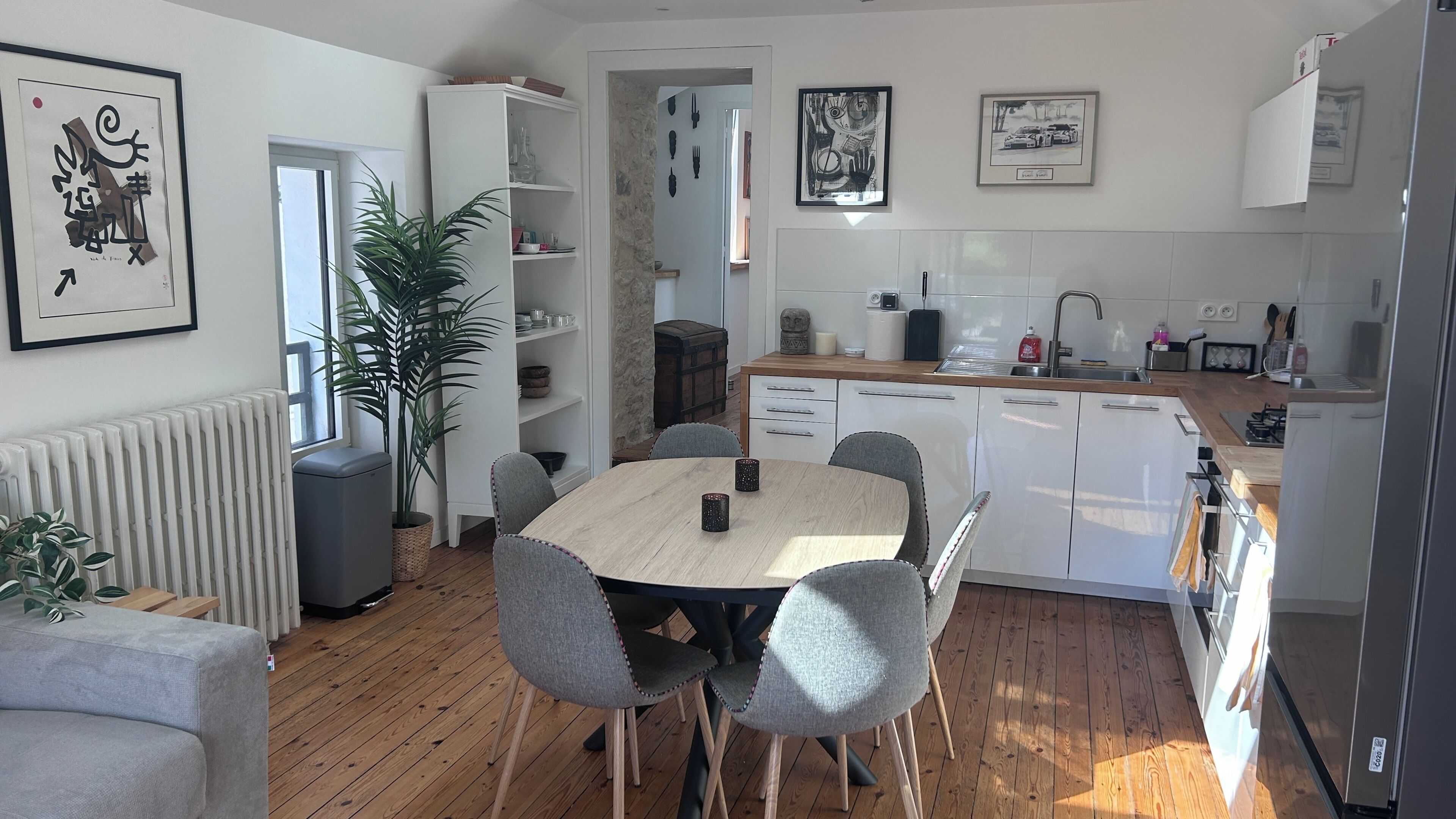 Une cuisine moderne avec des meubles blancs, une table à manger en bois, des chaises grises et une plante en pot.
