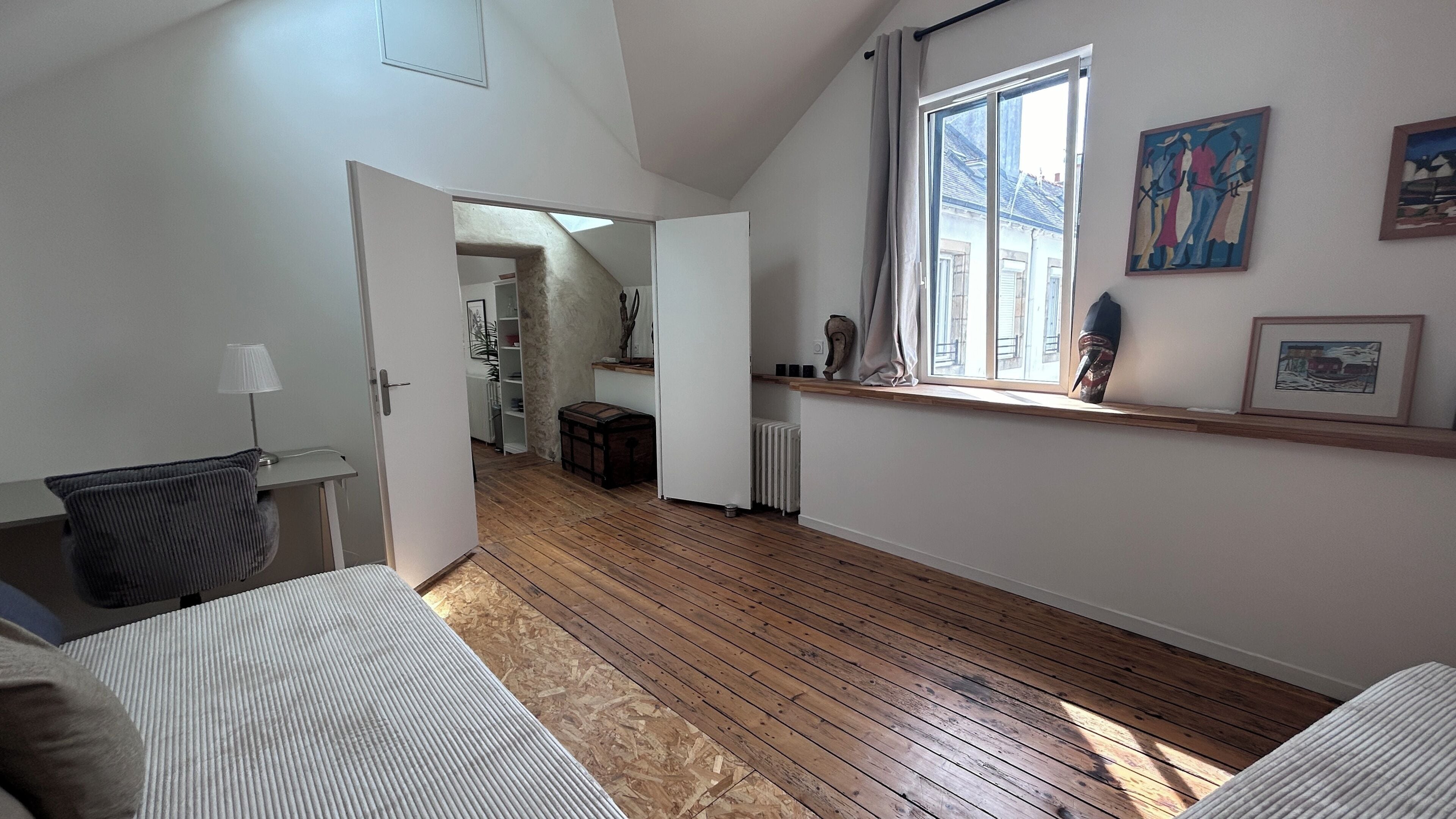 Une pièce avec un plancher en bois, un lit, un bureau, un radiateur et une fenêtre décorée d’œuvres d’art.