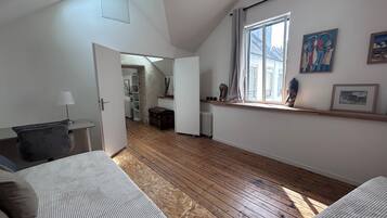 Une pièce avec un plancher en bois, un lit, un bureau, un radiateur et une fenêtre décorée d’œuvres d’art.