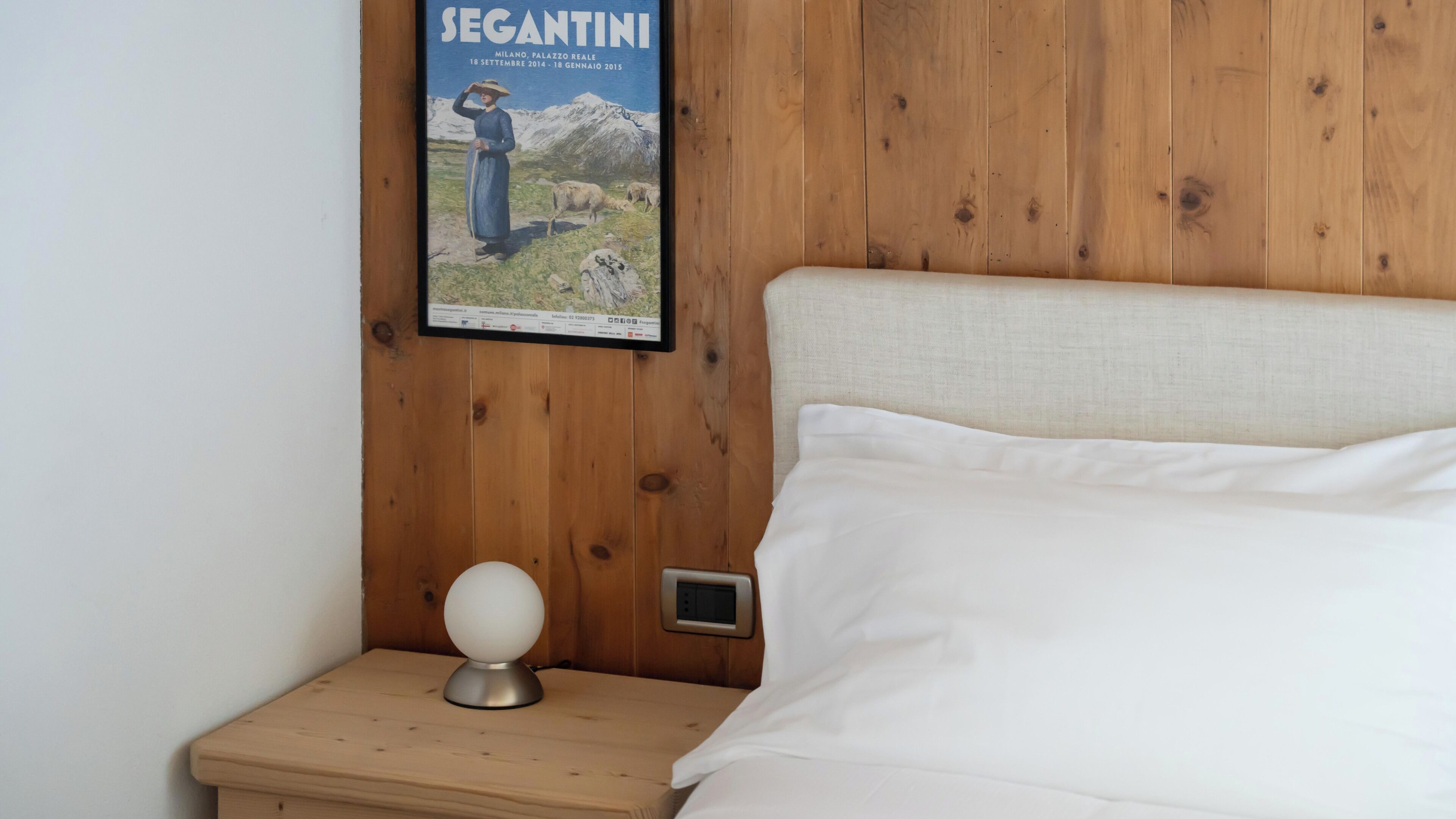 A bedroom with a wooden headboard, a poster on the wall, a bedside table with a lamp, and a bed with white linens.