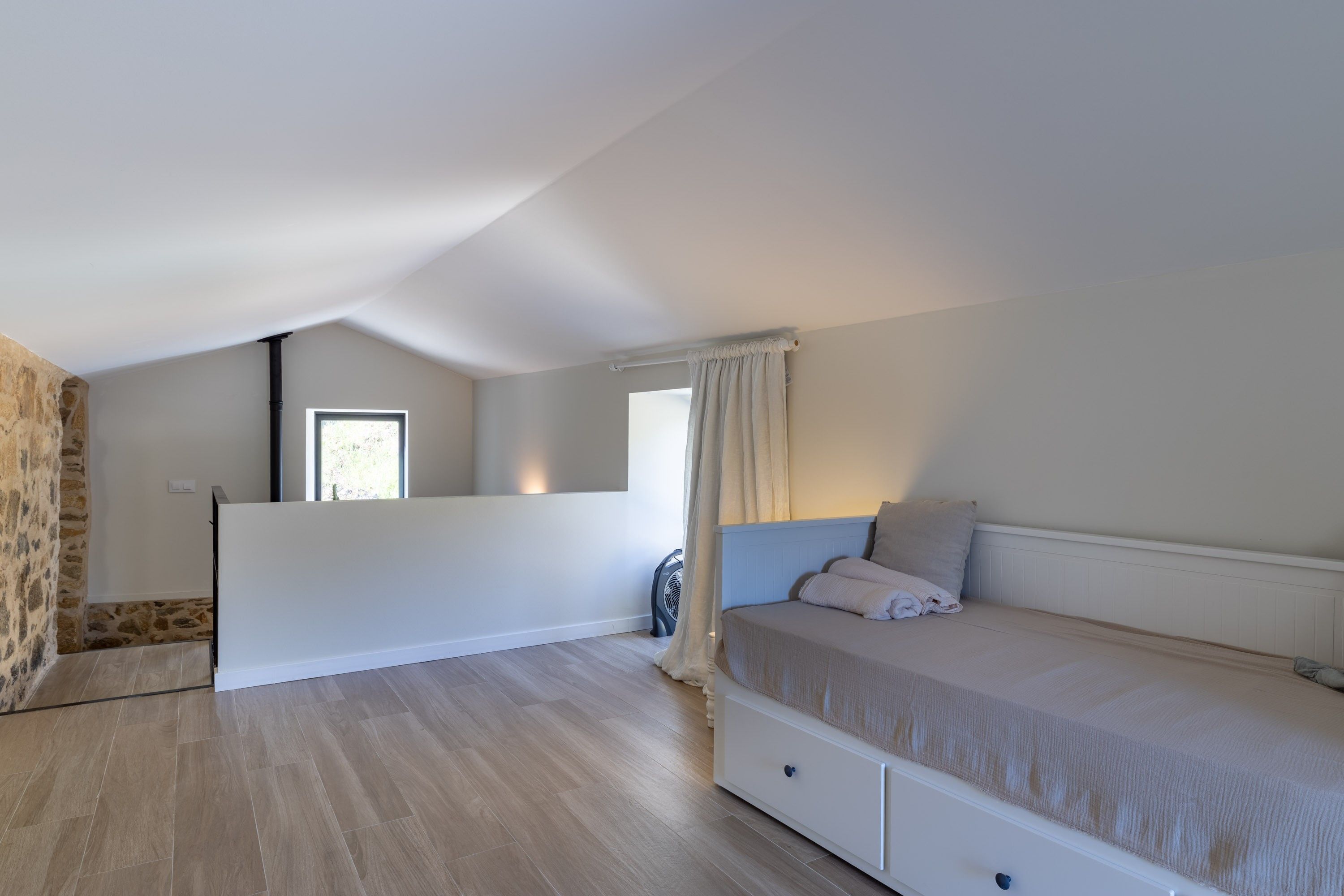 Une chambre avec un lit, une fenêtre, un ventilateur et un mur en pierre.