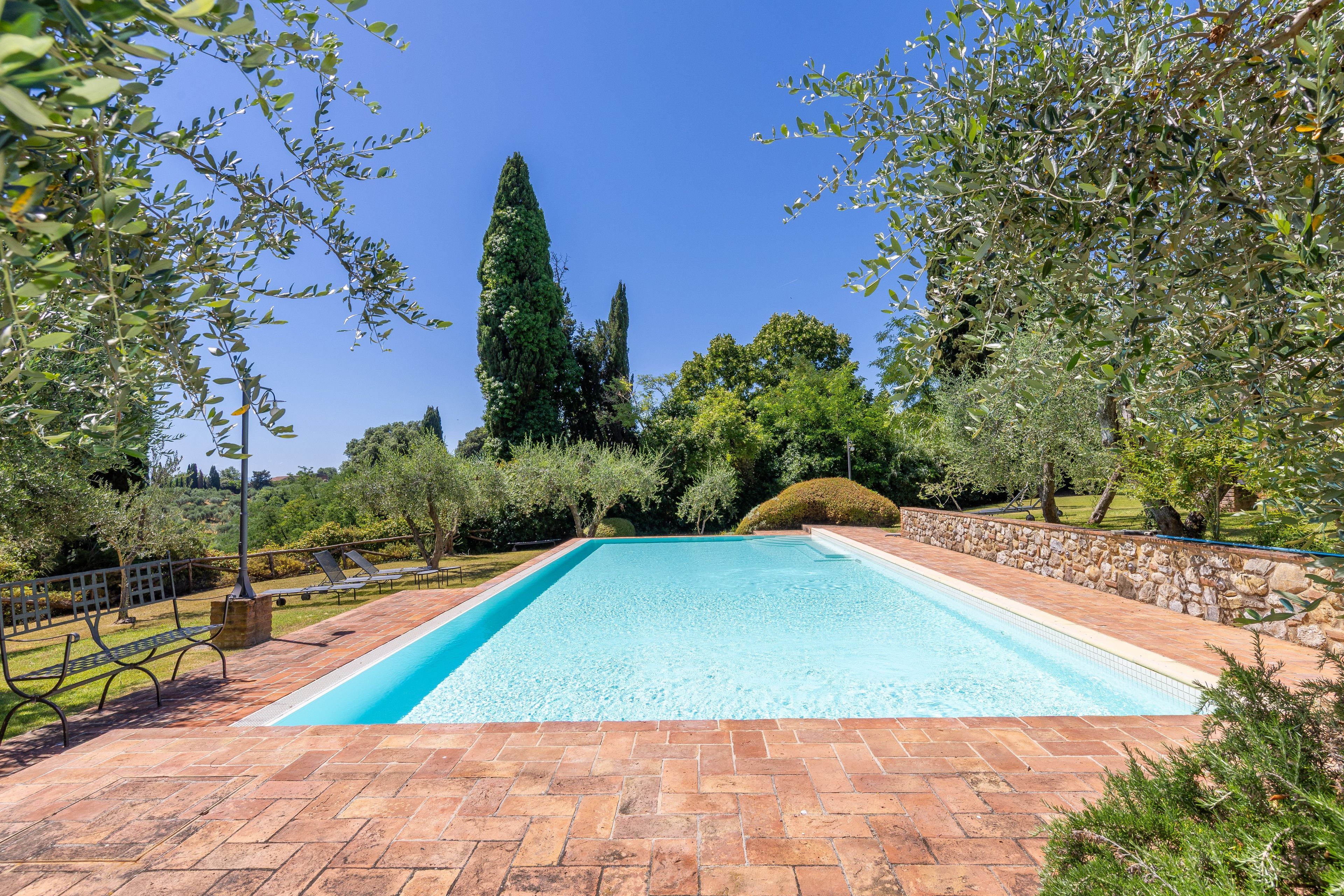 Une piscine rectangulaire bordée de pierres, entourée d’une terrasse en briques et avec des arbres en arrière-plan.