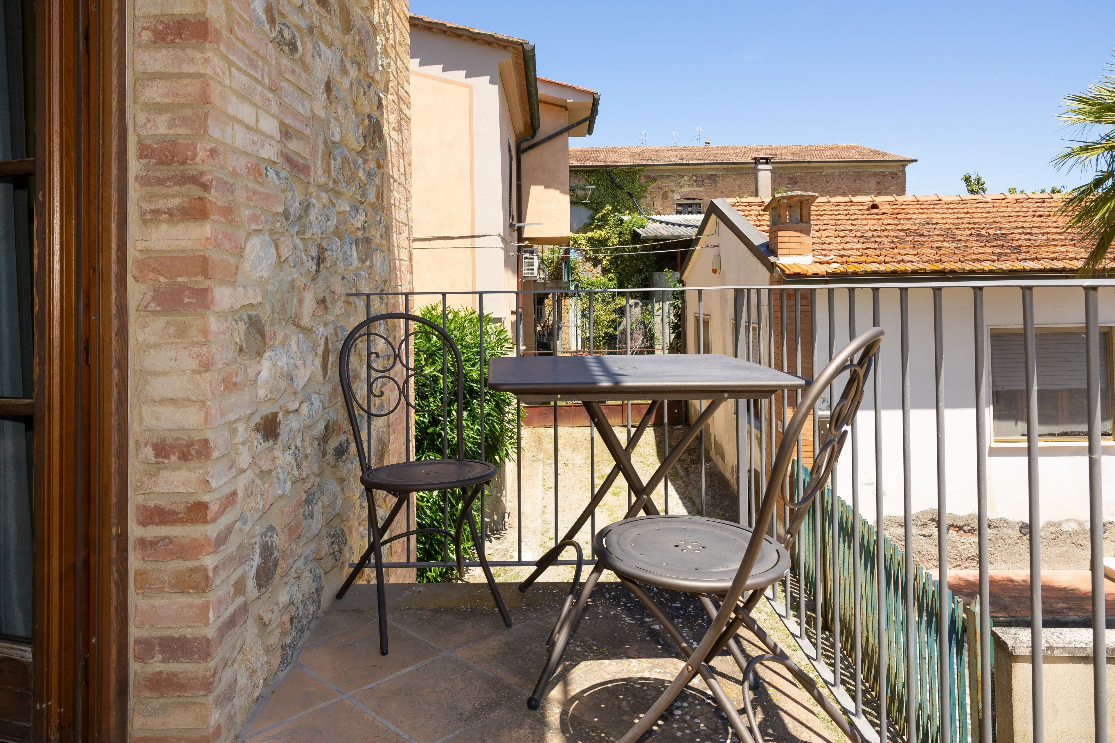 A balcony with a metal table and two chairs, a brick wall, and a tiled roof in the background.