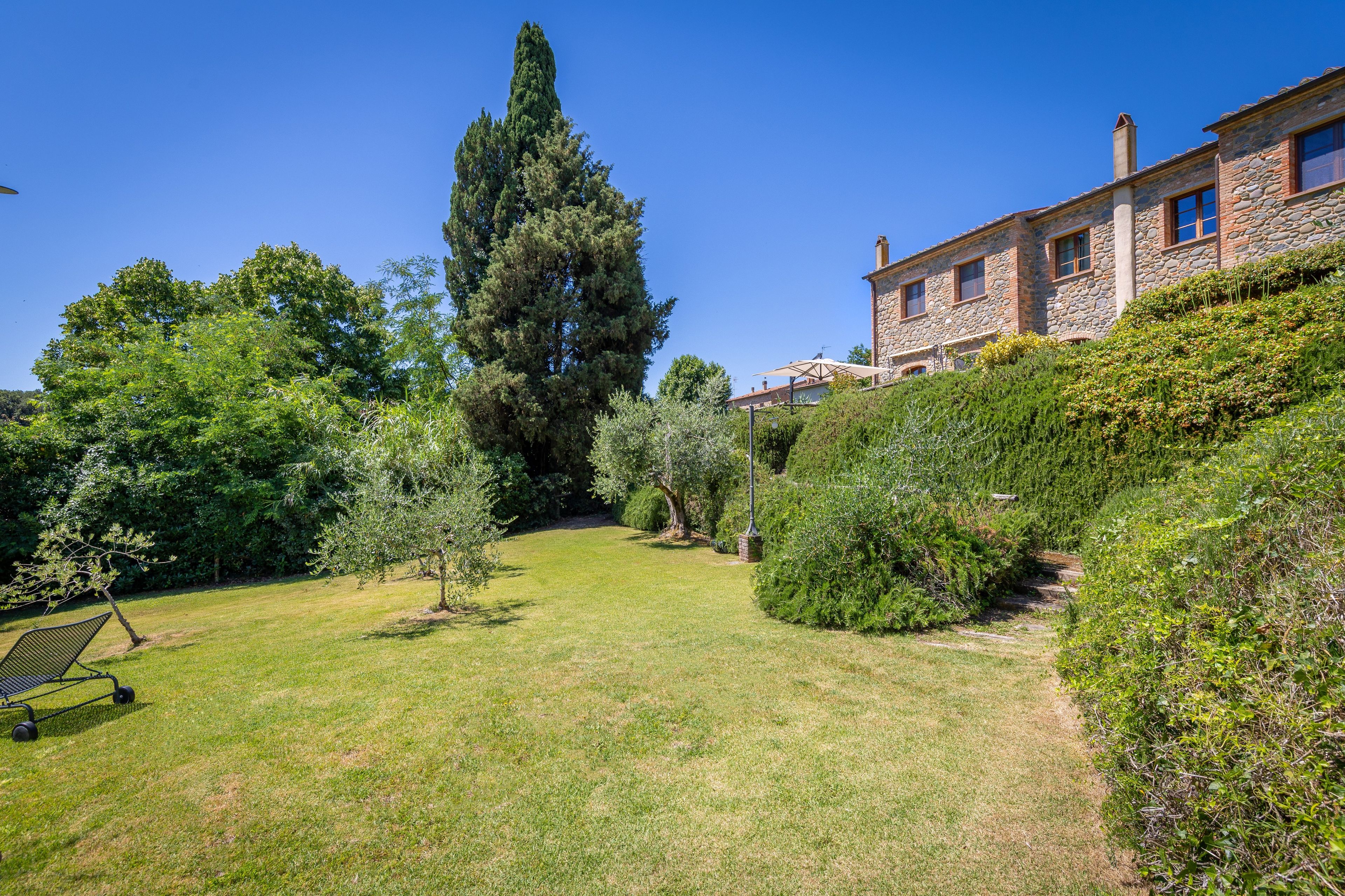 Un jardin bien entretenu, agrémenté de divers arbres et arbustes, un chemin dégagé et un bâtiment en pierre doté de nombreuses fenêtres.
