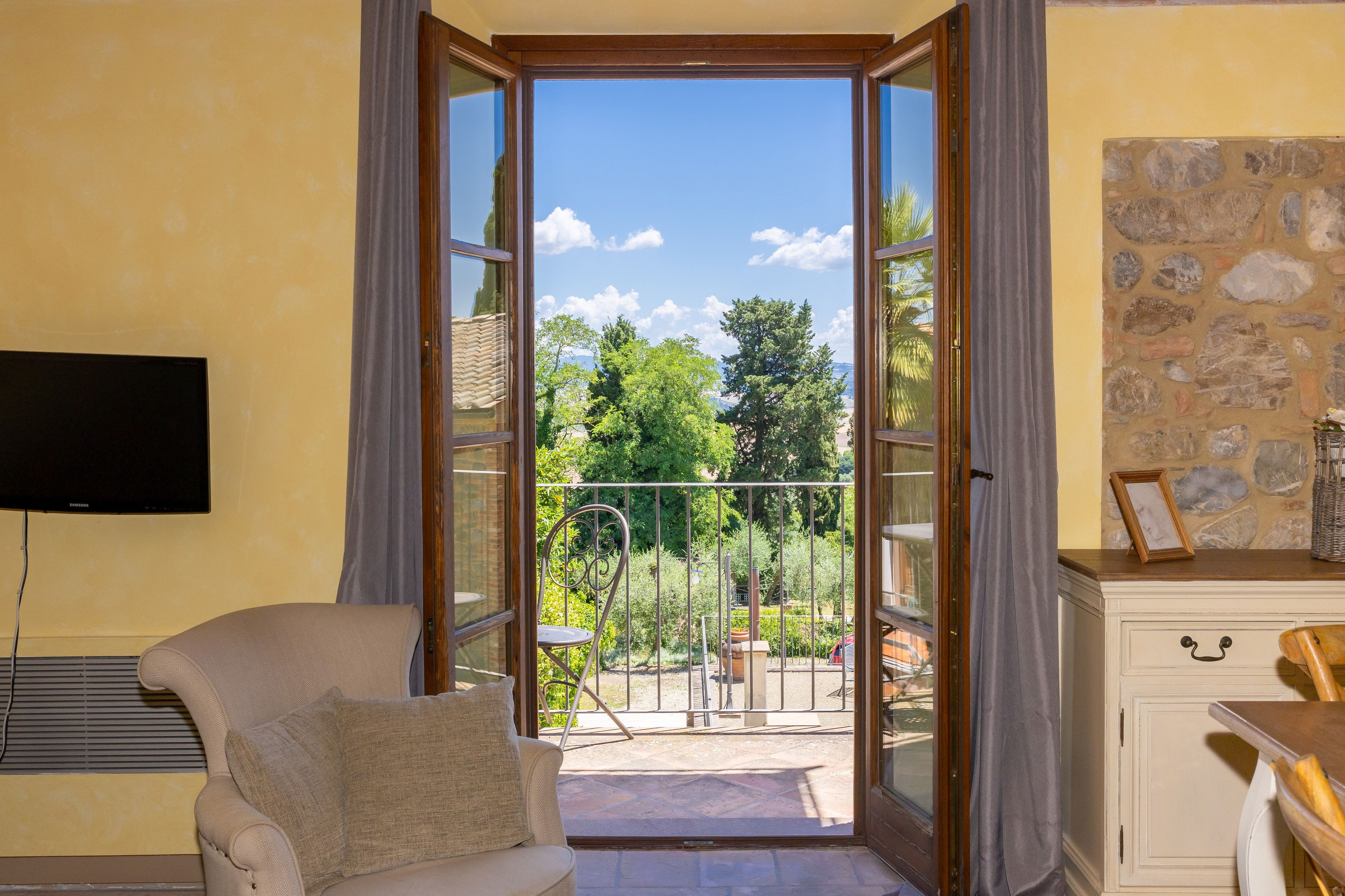 A room with a sofa, a television, a stone wall, and a balcony with a view of trees and a clear sky.