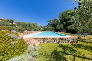 A rectangular swimming pool with a stone border, surrounded by well-maintained grass and various shrubs.