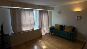 Een kamer met een houten vloer, een groene bank met kussens, een radiator en een raam met gordijnen.