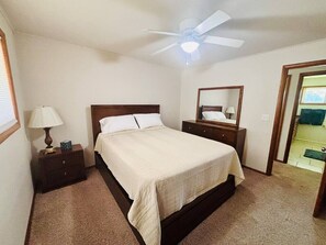 Une chambre à coucher comprenant un lit, des tables de chevet, un ventilateur de plafond, un miroir et une porte donnant sur une autre pièce.
