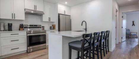 A modern kitchen with white cabinetry, a stainless steel oven, a refrigerator, and a bar area with stools.