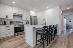 A modern kitchen with white cabinetry, a stainless steel oven, a refrigerator, and a bar area with stools. - The Point Lodge - Cozy Invermere Getaway (Invermere)