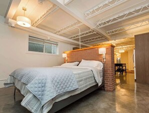 A modern bedroom with a bed, a brick accent wall, a ceiling with exposed beams, and a hallway leading to another room. - The City Haven | Sleeps 4 | (Calgary)