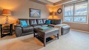 A living room with a sectional sofa, a coffee table, two lamps, a clock, and a window with a view.