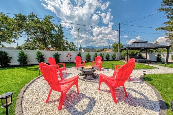 A circular outdoor seating area with a fire pit, surrounded by red chairs, set on a gravel surface with a white wall and greenery in the background. - Backyard Oasis - Hot Tub, BBQ, Outdoor Bar (Invermere)