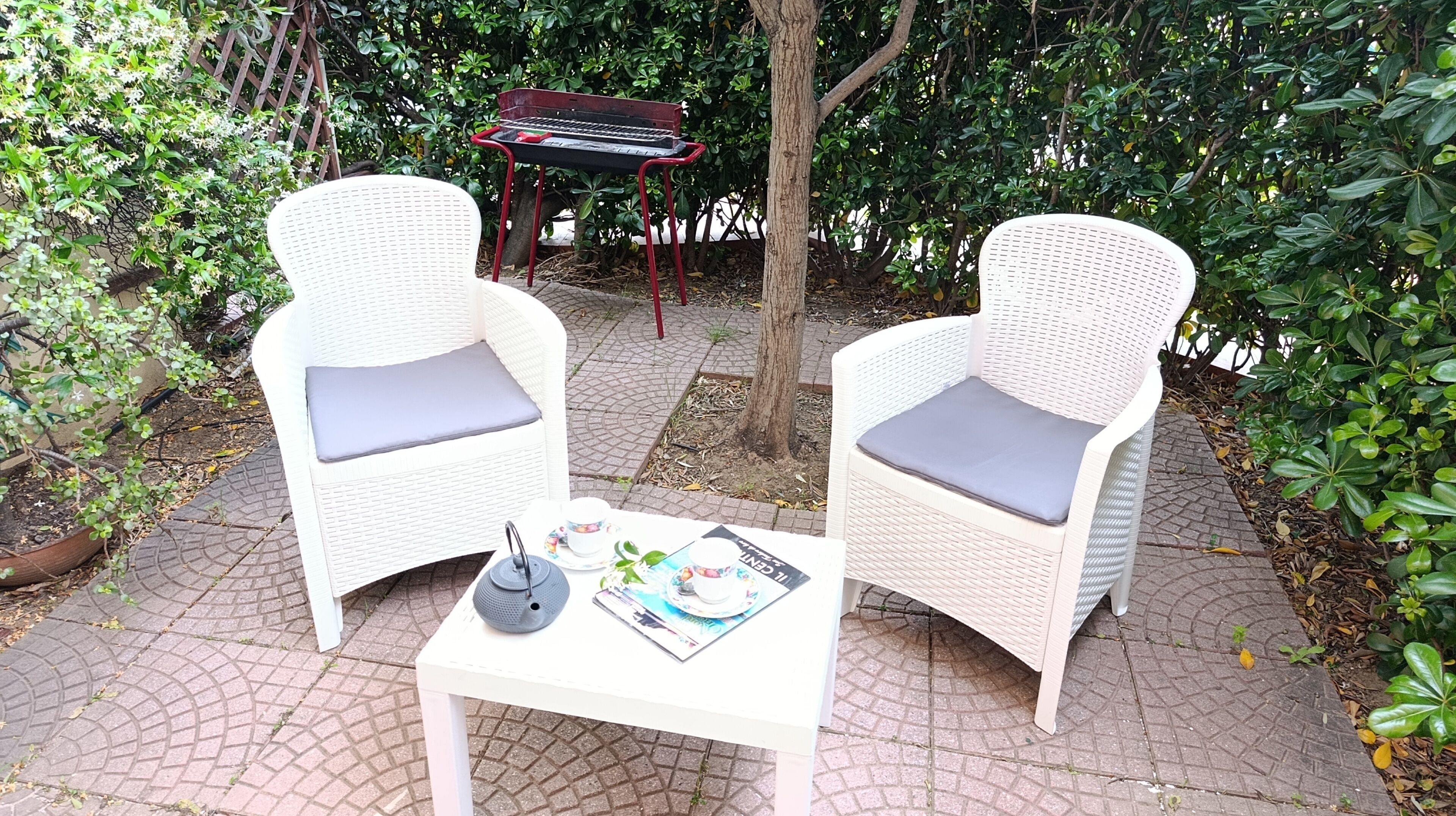 Two wicker chairs with grey cushions, a small table with a teapot, cups, and a magazine, and a red metal table with a keyboard in a garden setting.