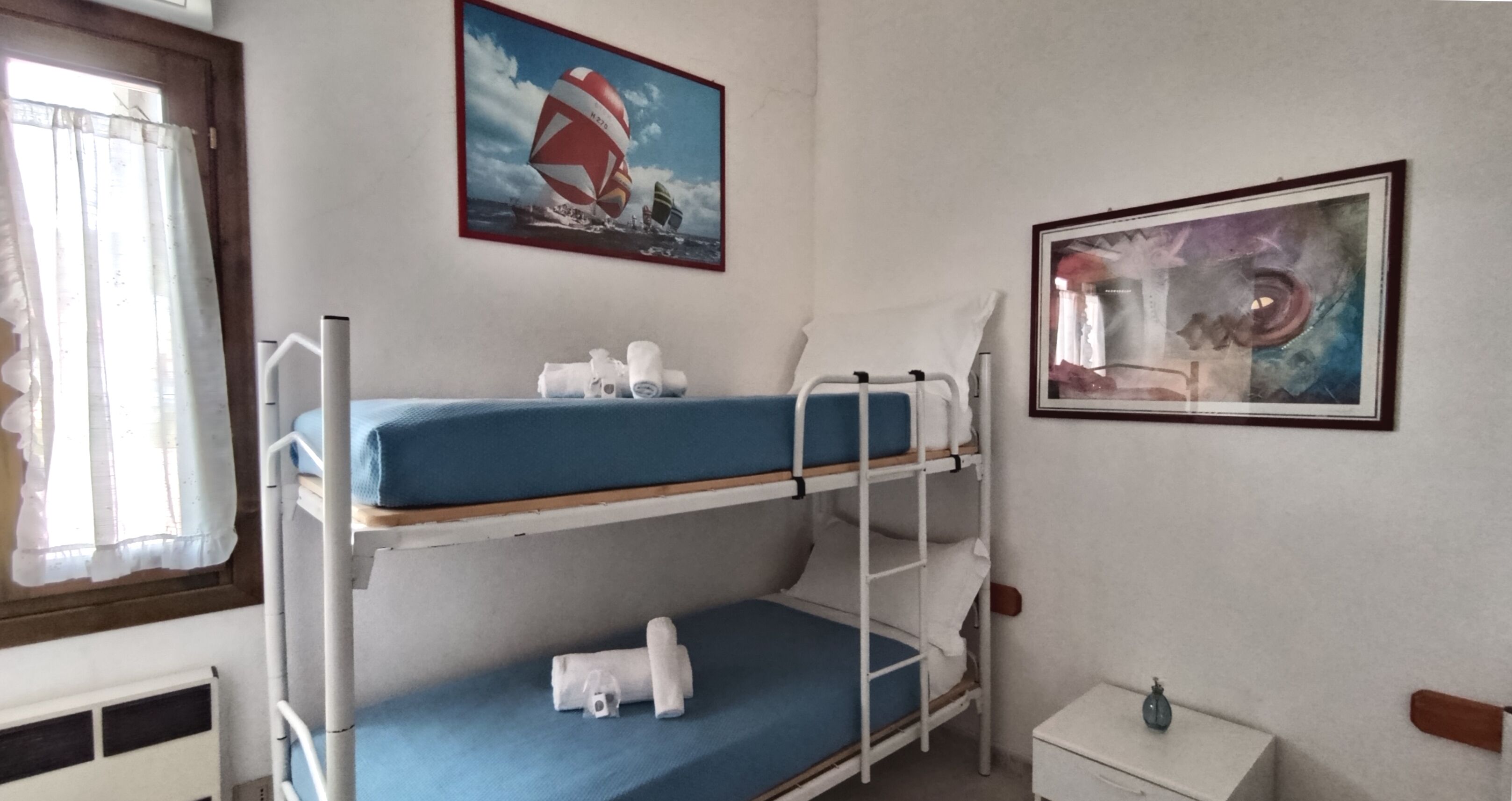 Een slaapkamer met stapelbed, een raam, een schilderij van een zeilboot en een klein nachtkastje.