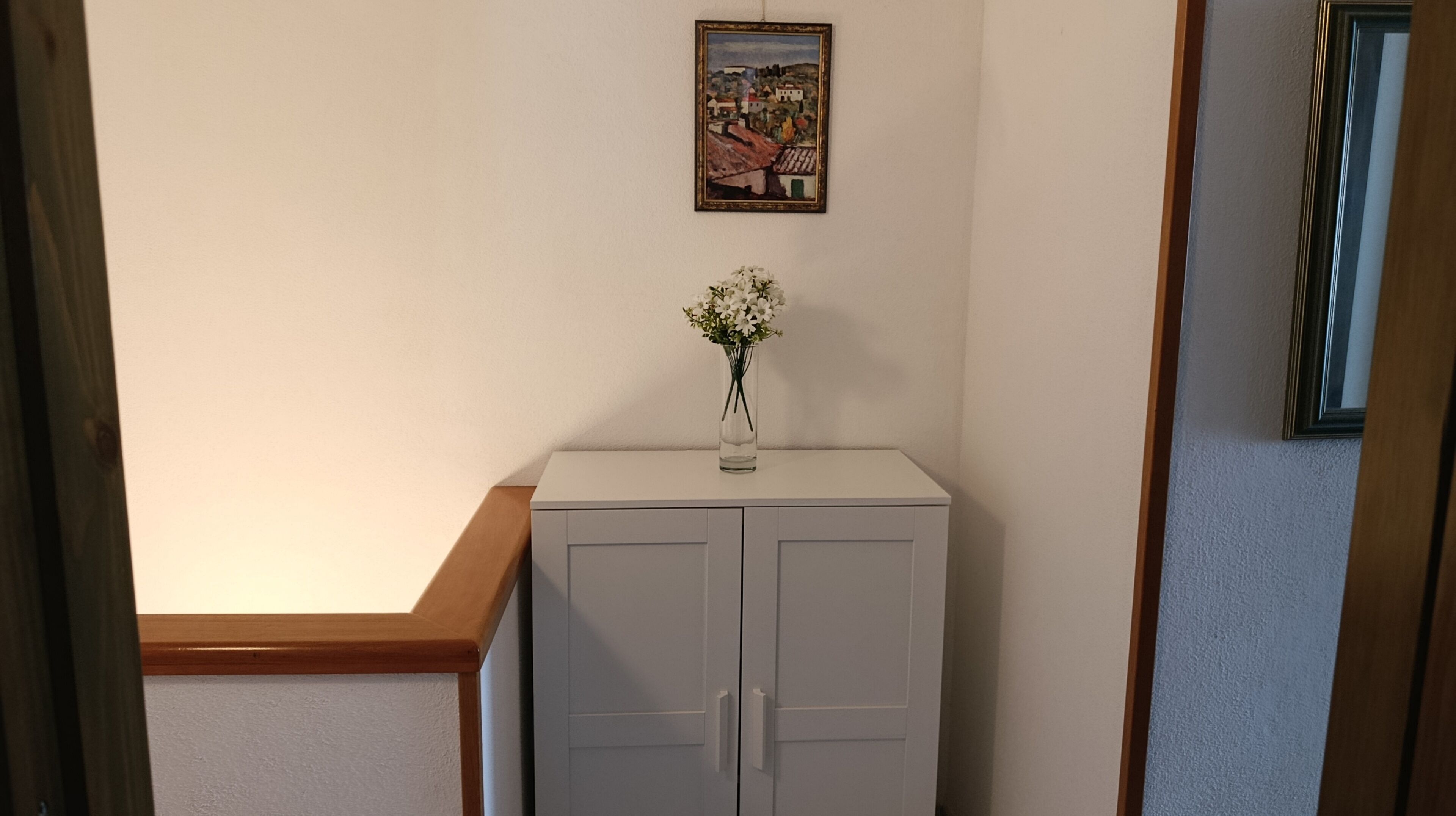 Un corridoio con un armadietto bianco, un vaso con fiori bianchi e un quadro appeso al muro.
