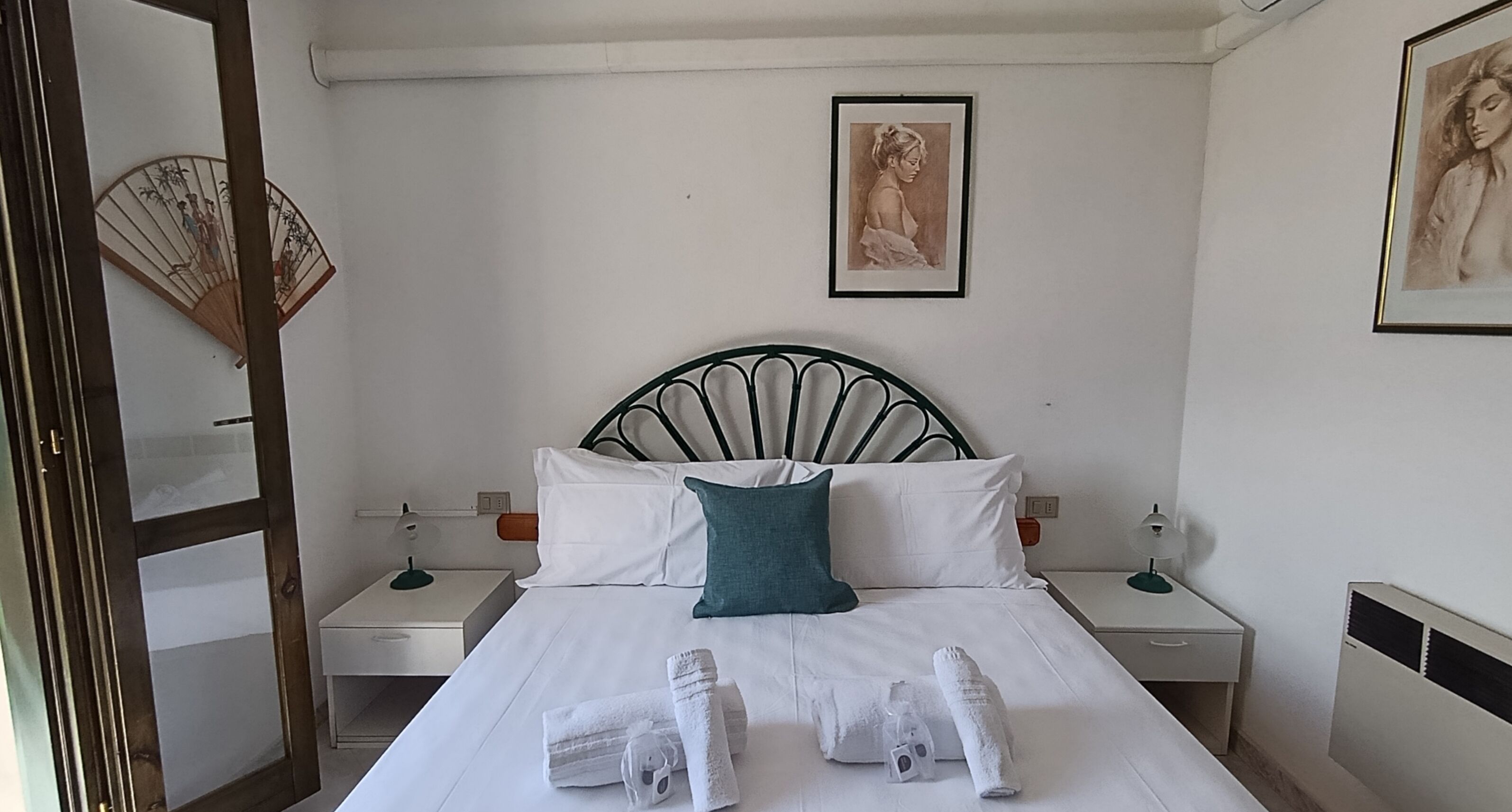 Un letto rifatto con lenzuola bianche e un cuscino color petrolio, due comodini con lampade, un quadro incorniciato al muro e una ventola tradizionale.
