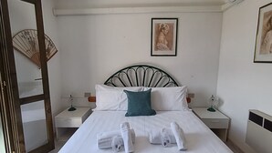 Una cama bien hecha con sábanas blancas y un almohadón verde azulado, dos mesitas de noche con lámparas, un cuadro en la pared y un abanico tradicional.