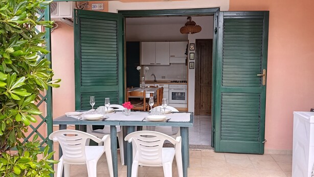 Una puerta abierta que da a un comedor con una mesa dispuesta para dos, sillas y una cocina al fondo.