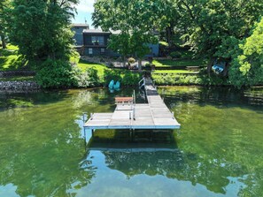 A wooden dock extending into a calm body of water, surrounded by lush greenery and a residential building in the background.