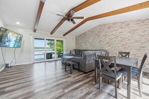 A spacious living area with a wooden ceiling, a gray sofa, a dining table with chairs, and a TV on the wall.