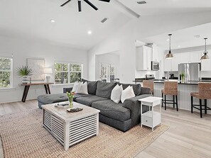 Uma sala de estar espaçosa com um sofá modular cinza, uma mesa de centro e uma área de cozinha com armários brancos e um banquinho.