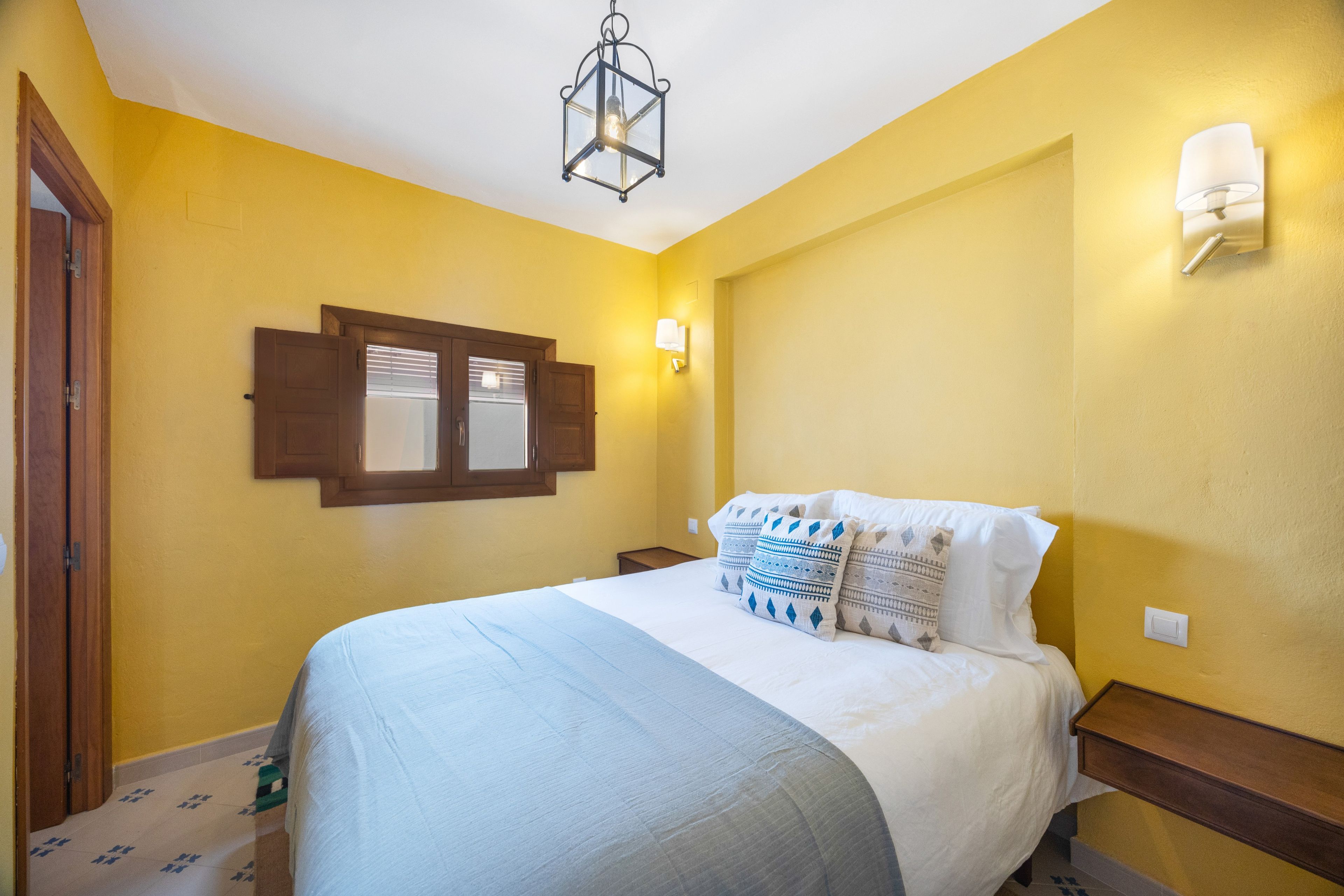 A bedroom with a bed, a window with shutters, a wall-mounted light, and a door.
