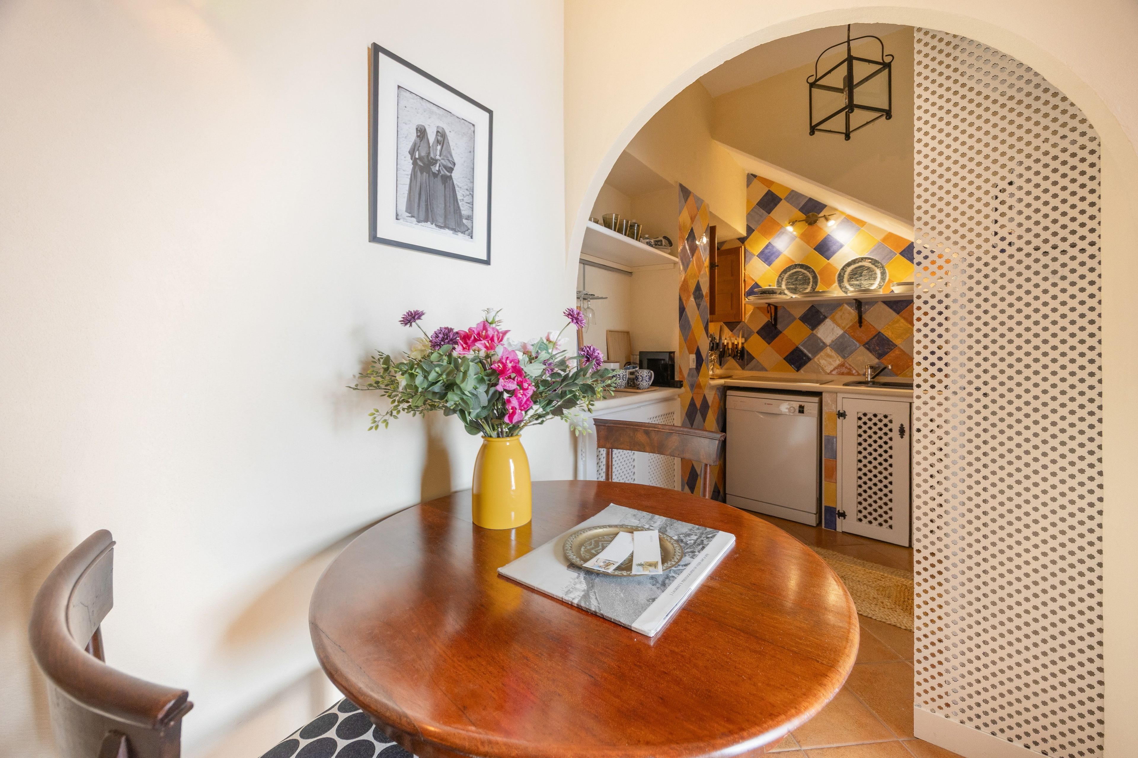 A dining area with a round wooden table, a yellow vase with flowers, and a framed picture on the wall.