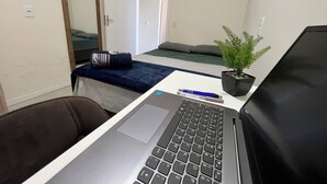 A small room with a bed, a desk with a laptop, a chair, a potted plant, and a door. - Casa piscina praia da pinheira  (Palhoça)
