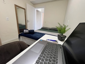 A small room with a bed, a desk with a laptop, a chair, a potted plant, and a door. - Casa piscina praia da pinheira (Palhoça)