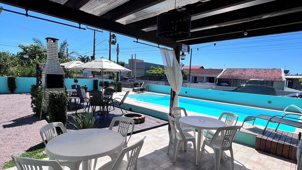 An outdoor patio with a pool, white plastic chairs, and a brick oven. - Casa piscina praia da pinheira  (Palhoça)