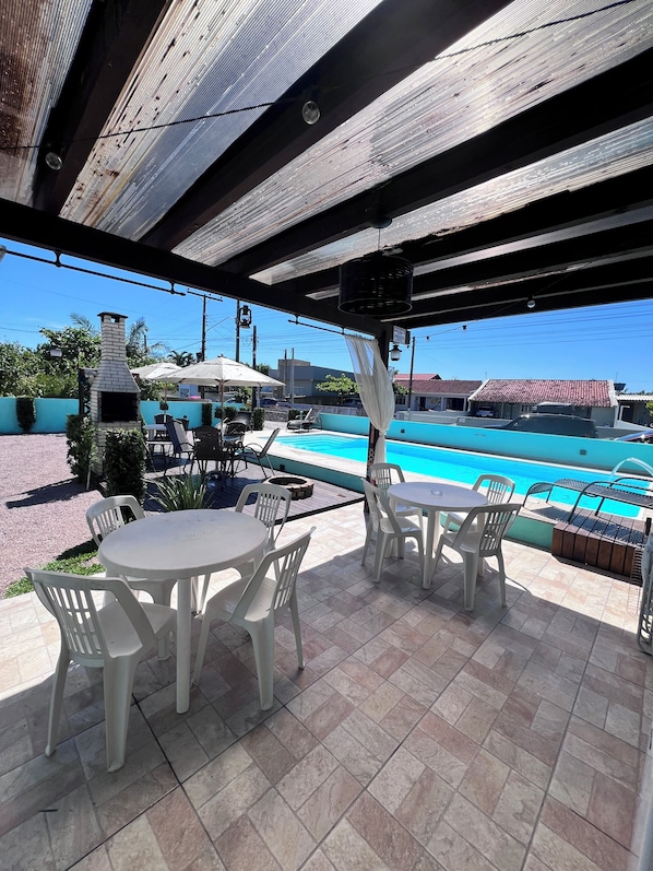 An outdoor patio with a pool, white plastic chairs, and a brick oven. - Casa piscina praia da pinheira (Palhoça)