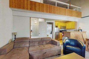 Ein Wohnzimmer mit einer Couch, einem Sessel, einem Esstisch und einer Küchenzeile mit gelben Schränken und einer Mikrowelle.
