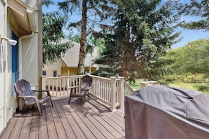Eine Holzterrasse mit zwei Adirondack-Stühlen, einem Grill und einem Haus im Hintergrund.
