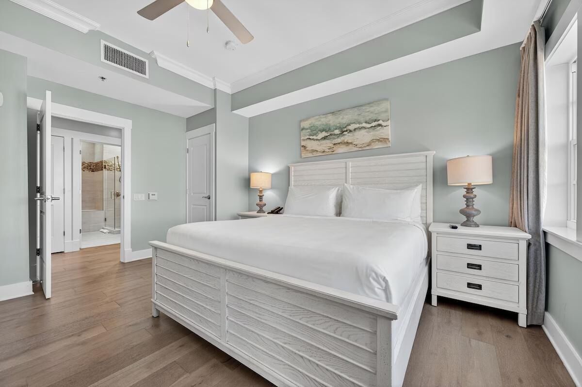A bedroom with a large bed, two bedside tables with lamps, a ceiling fan, and a painting on the wall.