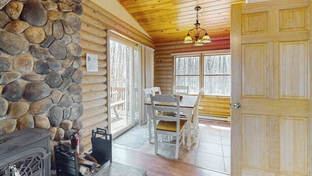 A log cabin interior with a stone wall, wooden ceiling, a dining table with chairs, and a fireplace.