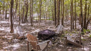A campsite with four wooden chairs arranged around a central fire pit, surrounded by trees.