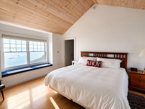 Ein Schlafzimmer mit großem Fenster, einem Bett mit weißer Bettwäsche, einem Holz-Kopfteil und einem Nachttisch mit Lampe.