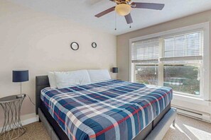 Ein Schlafzimmer mit einem Bett, einem Deckenventilator, einem Fenster mit Jalousien und einem kleinen Tisch mit einer Lampe.