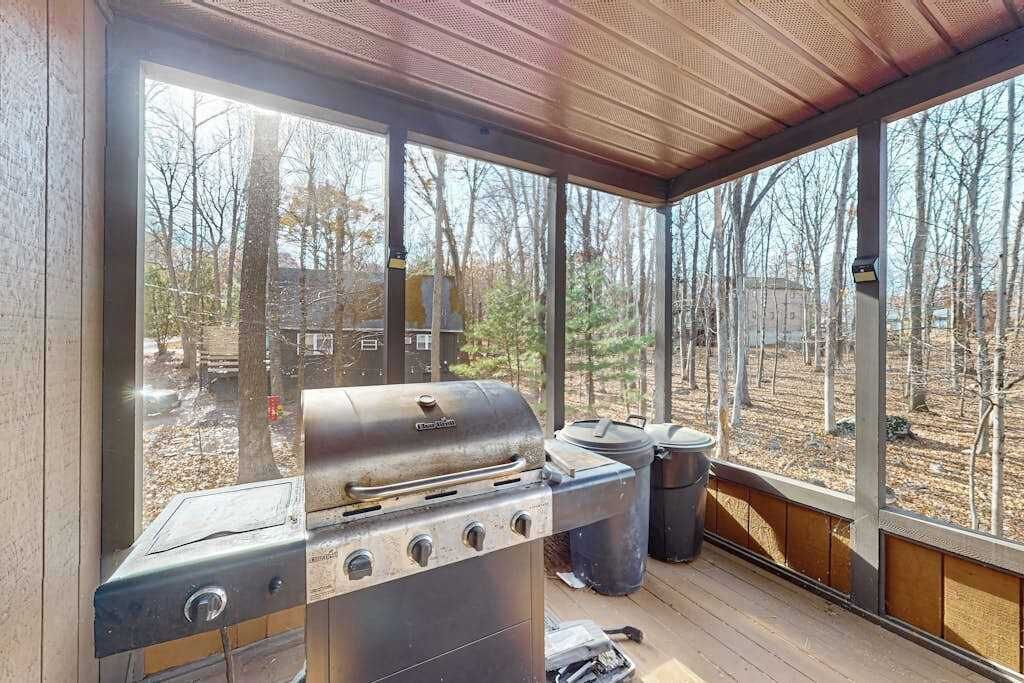Ein überdachter Außenbereich mit Grill, Müllbehältern und Blick auf Bäume und ein Gebäude.