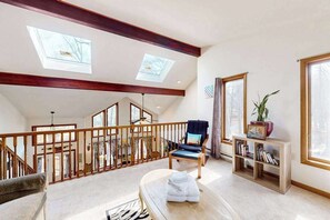 Un loft spacieux avec de hauts plafonds, des fenêtres de toit, un escalier en bois, un coin salon avec un fauteuil et une petite table, ainsi qu’une bibliothèque.
