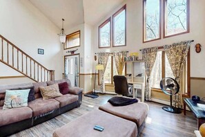 A spacious living room with a staircase, a sofa, ottomans, a desk, and large windows. - 3BR Spacious Dog-Friendly House (East Stroudsburg)