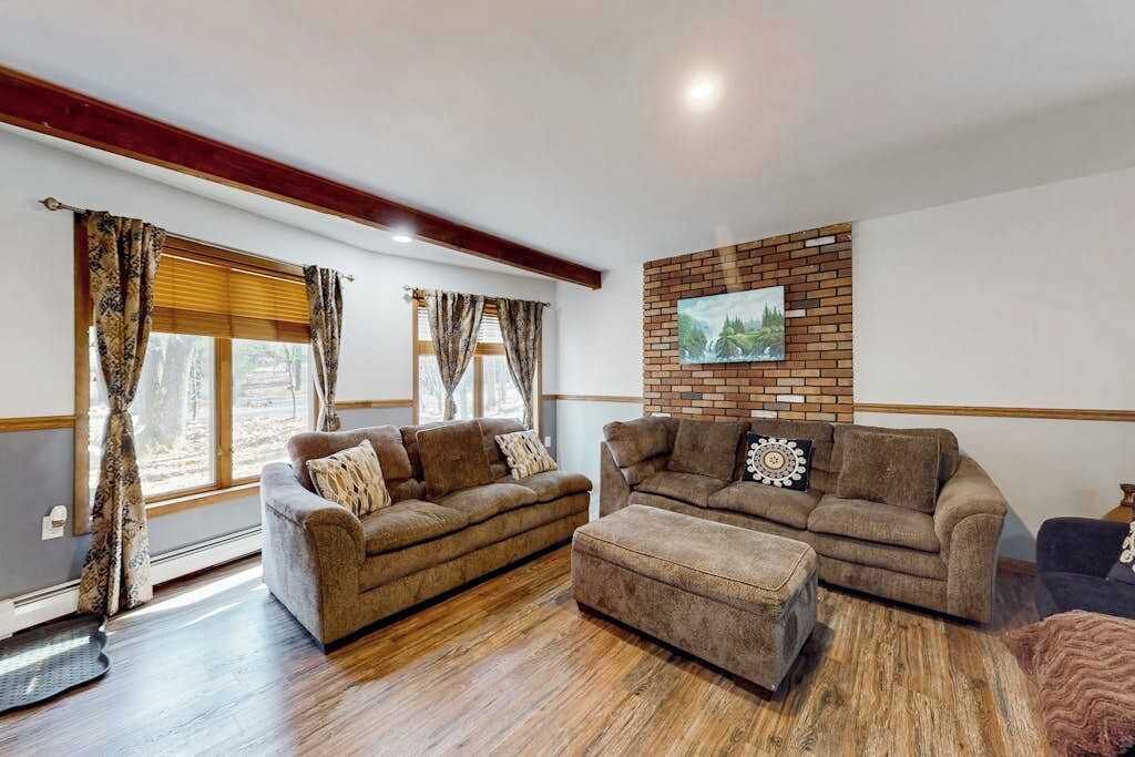 Una sala de estar con dos sofás y una mesa de centro, piso de madera, pared de ladrillo decorativa y una ventana con cortinas.
