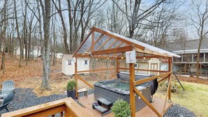 A hot tub under a wooden pergola with a deck, surrounded by trees and a house in the background. - Lakeside Chalet with Hot Tub, Fire Pit & Game Room (Blakeslee)